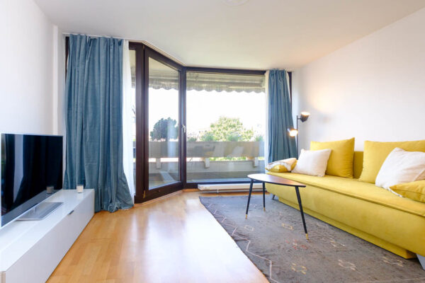 Wohnzimmer Blick auf gelbes Schlafsofa, Couchtisch, TV, bodentiefe Fenster mit Zugang zum Balkon