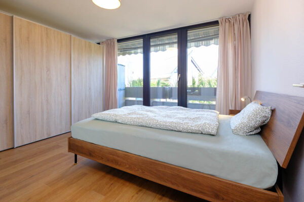 Blick von der Schlafzimmertüre auf das 140cm Bett, den grossen Schiebetürenschrank und die Bodentiefen Fenster zum Balkon